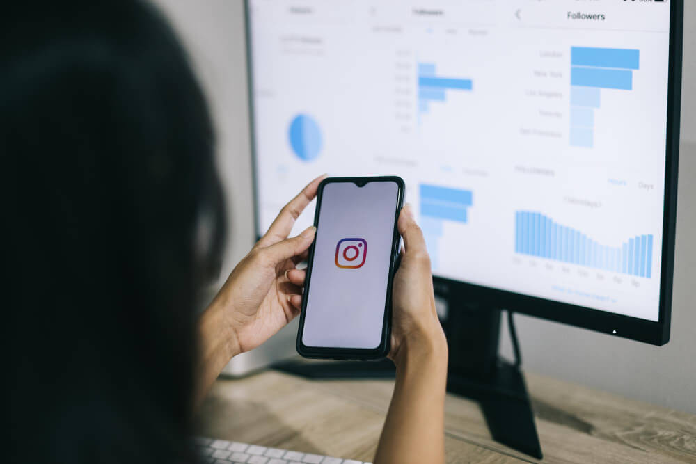 mãos segurando fone com a logo do instagram e ao fundo tela de computador mostrando gráficos de SEO